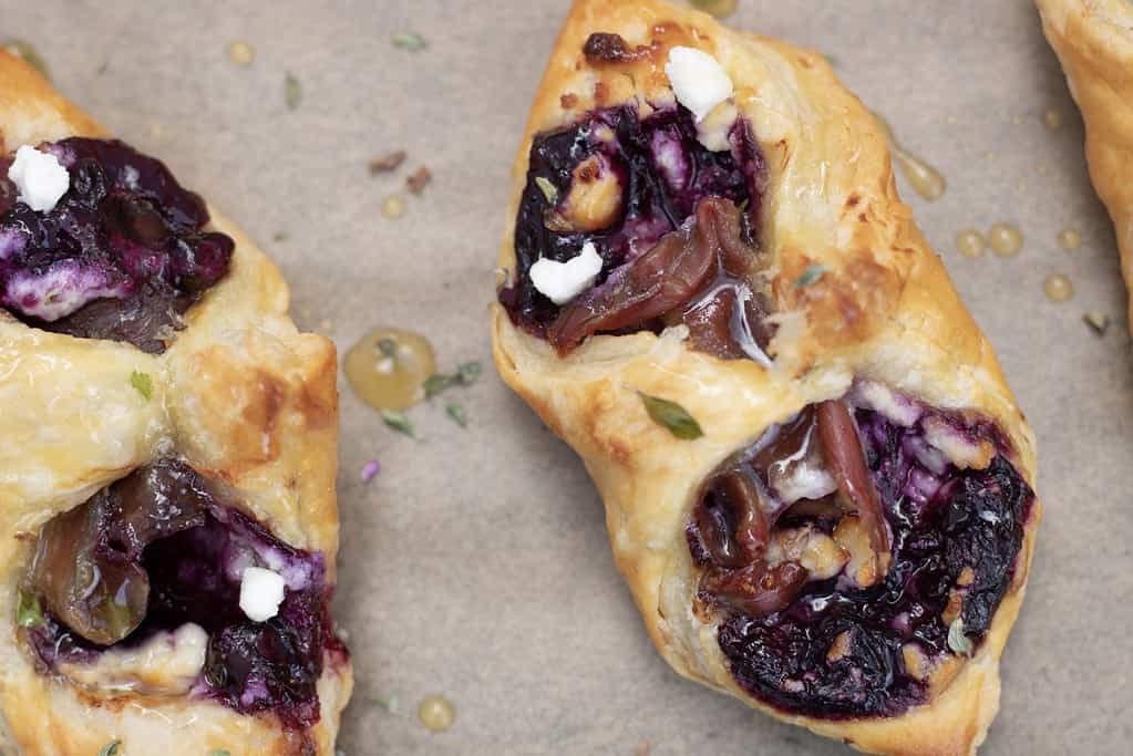A puff pastry square is filled with blueberry filling, a slice of prosciutto and goat cheese crumbles. The two corners are folded over each other to create a pouch. It’s baked until golden and flaky and drizzled with honey thyme.