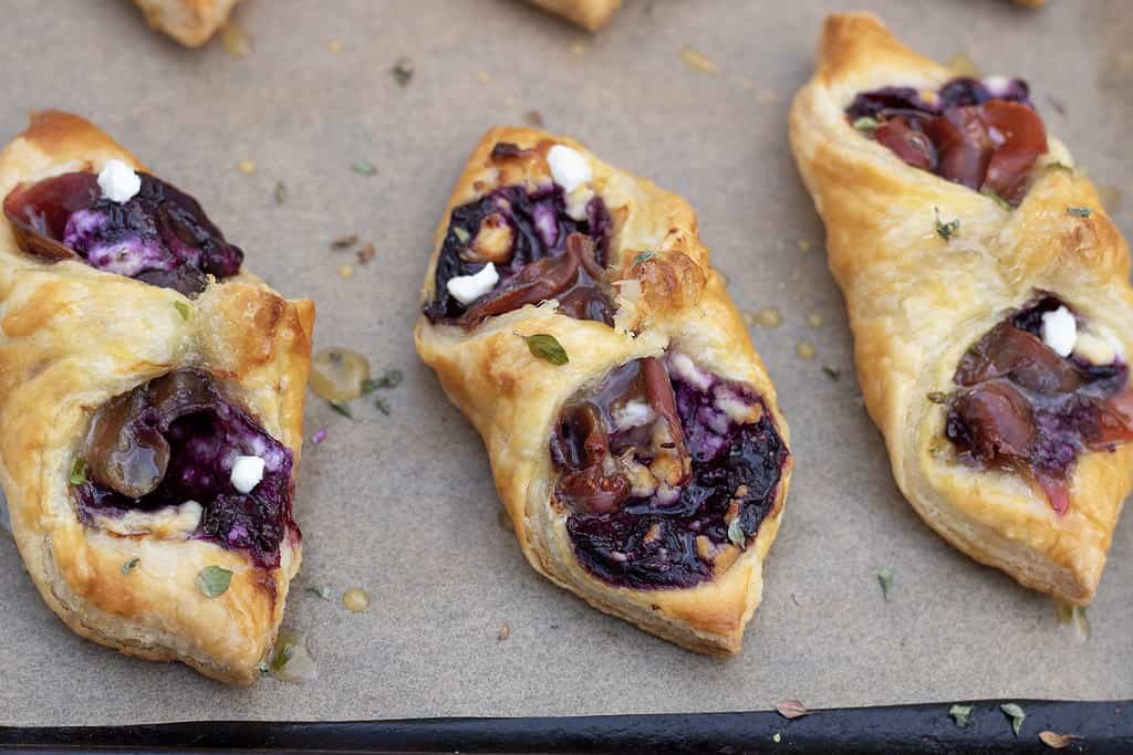 Puff pastry appetizers are golden and flaky. The inside is purple from the blueberry filling. The goat cheese crumbles are slightly browned and you can see the honey thyme dripping off the sides of the appetizers