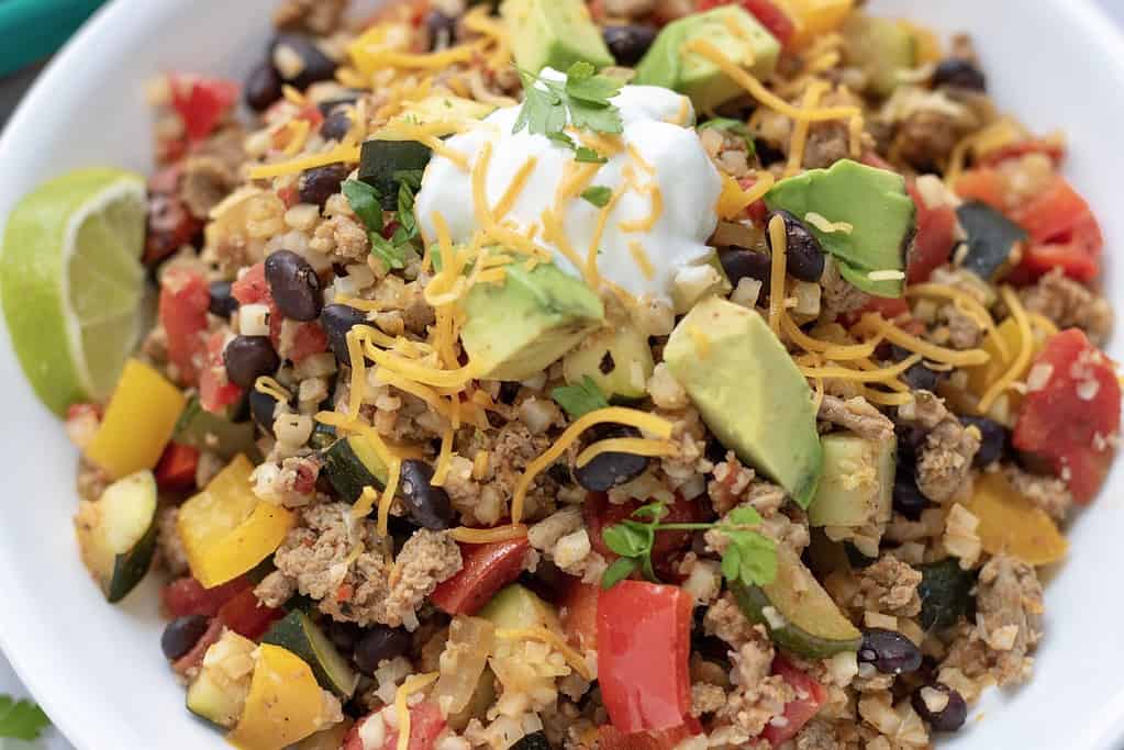 A large white bowl filled with cauliflower rice that's made in a skillet with ground turkey, black beans, peppers, fire roasted tomatoes and zucchini. It's topped with a spoonful of Greek yogurt and fresh cilantro and served with a piece of lime. It's a simple and easy healthy dinner recipe or great for low carb meal prep.