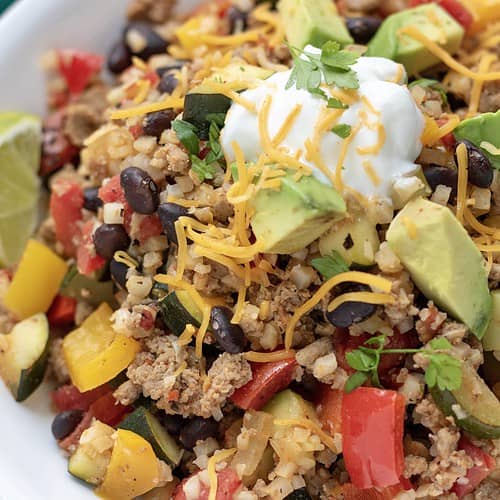 A large white bowl filled with cauliflower rice that's made in a skillet with ground turkey, black beans, peppers, fire roasted tomatoes and zucchini. It's topped with a spoonful of Greek yogurt and fresh cilantro and served with a piece of lime. It's a simple and easy healthy dinner recipe or great for low carb meal prep.
