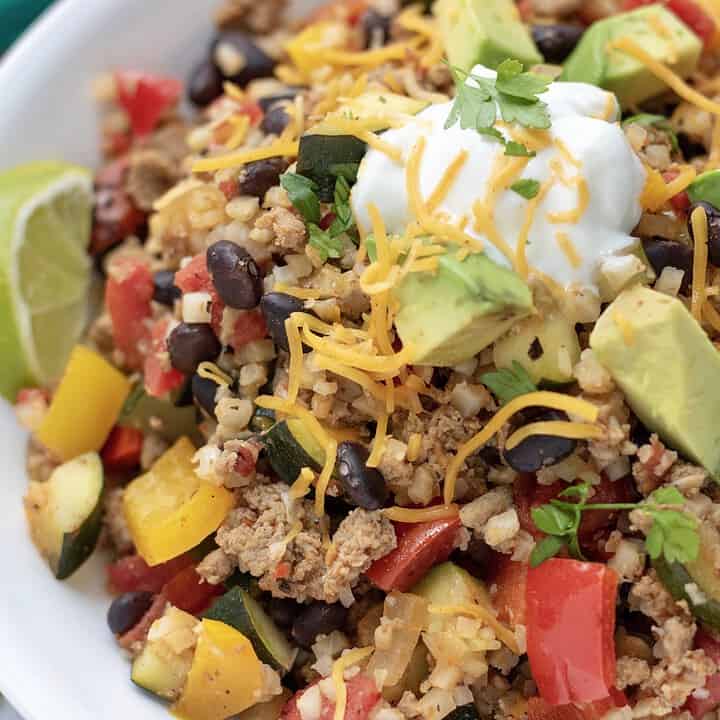 A large white bowl filled with cauliflower rice that's made in a skillet with ground turkey, black beans, peppers, fire roasted tomatoes and zucchini. It's topped with a spoonful of Greek yogurt and fresh cilantro and served with a piece of lime. It's a simple and easy healthy dinner recipe or great for low carb meal prep.