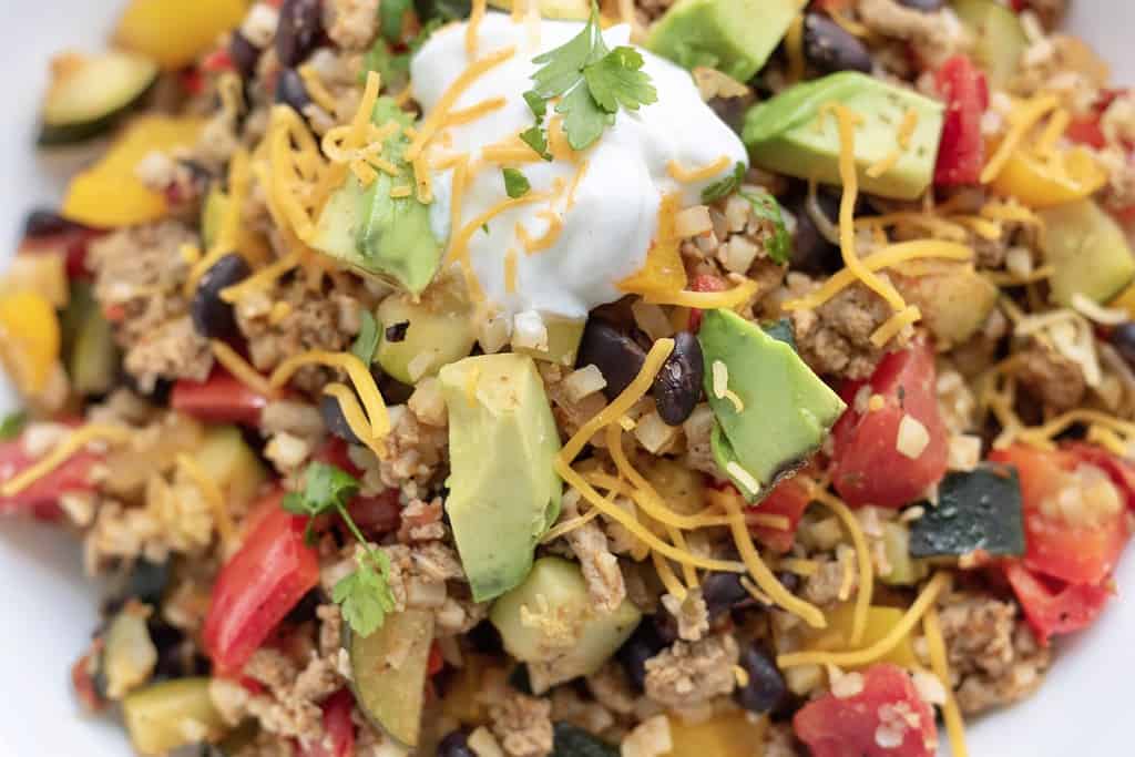 A bowl full of ground meat, black beans, zucchini, tomatoes and mexican cauliflower rice. It's topped with shredded cheese, a dollop of Greek yogurt and cilantro. It's a bright and flavorful low carb recipe that's perfect for meal prep.