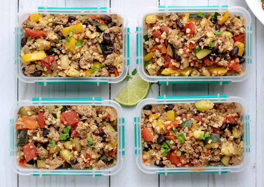 Four small containers packed with caulfilower rice that's made with mexican seasoning, zucchini, black beans, ground meat, cilantro and peppers. They're a great low carb meal prep recipe that's easy and healthy.