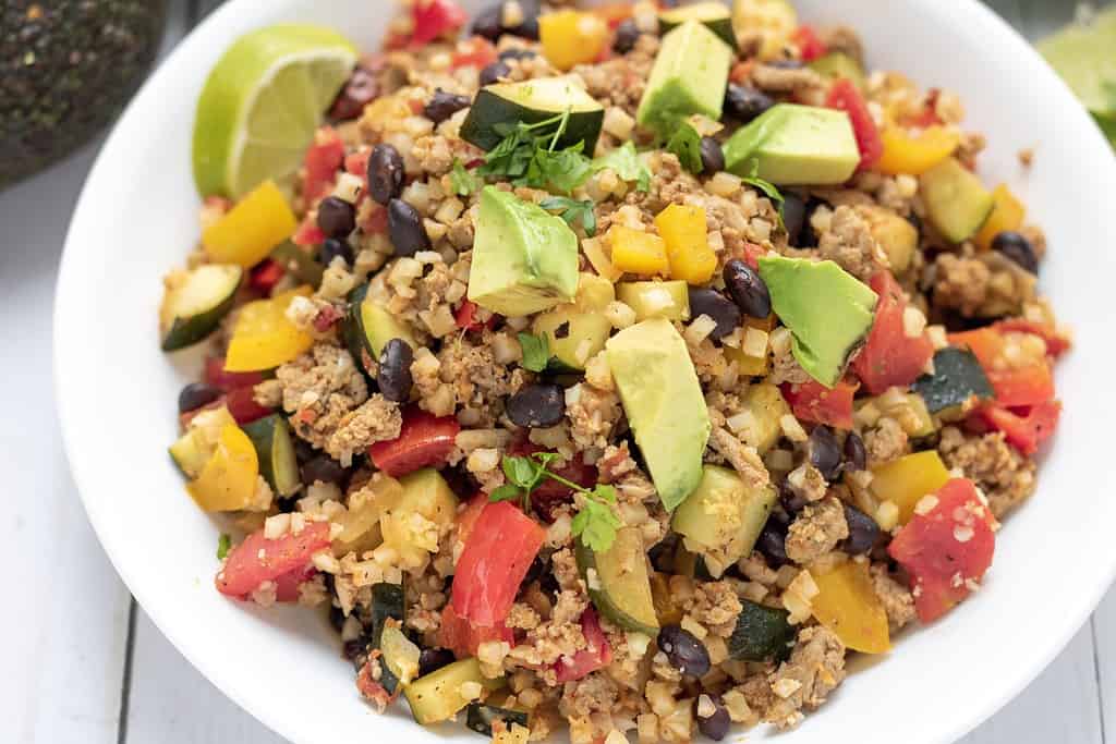 A white bowl full of cauliflower rice with black beans, ground turkey, peppers, roasted tomatoes, zucchini and mexican seasoning. It's a low carb and healthy dinner that's super easy to make!