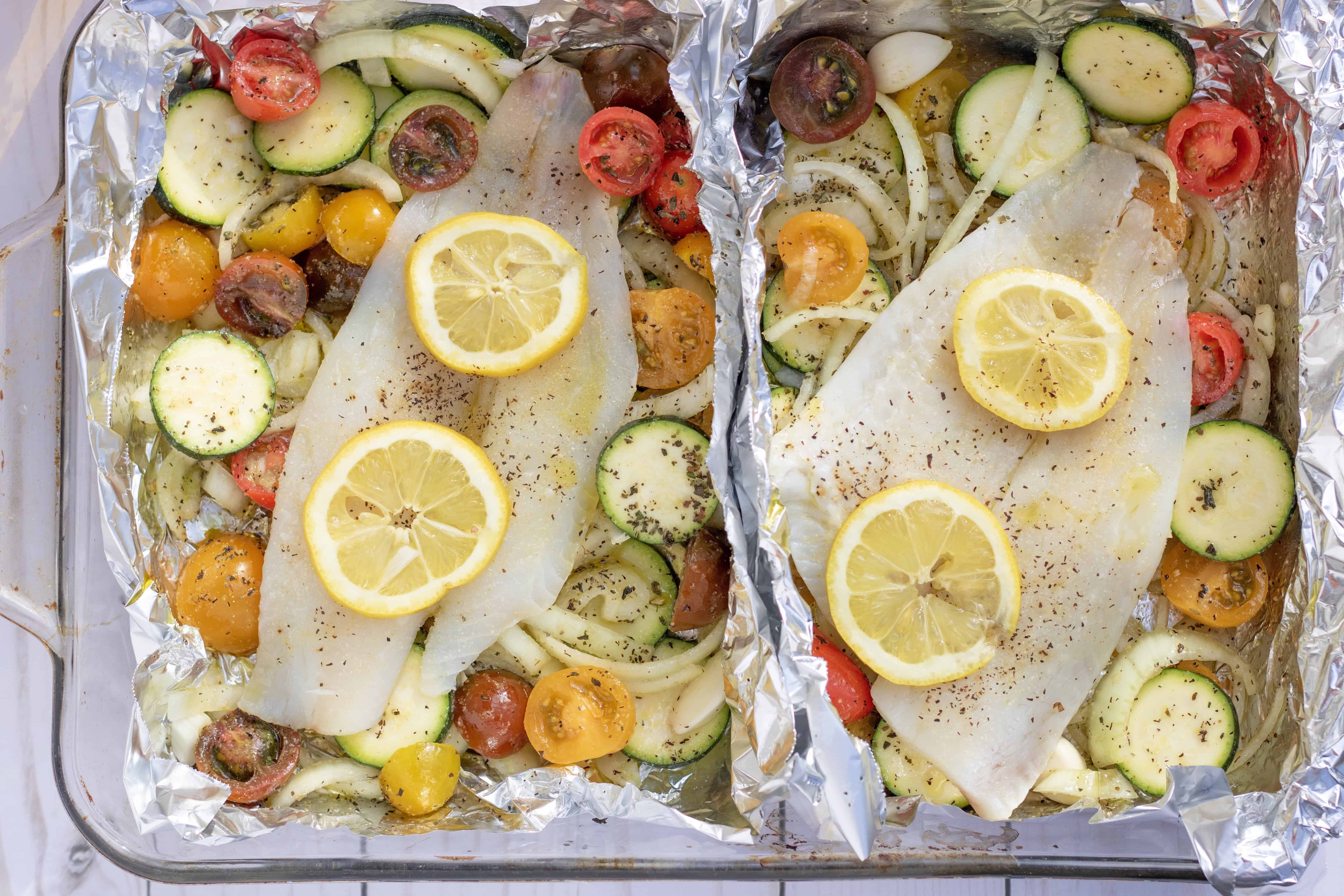 A glass 13x9” baking dish with two foil packets that are filled with vegetables and topped with white fish. It’s to be baked in the oven for an easy family dinner that’s healthy and quick to make. It’s got vibrant colorful cherry tomatoes, sliced zucchini and onions.