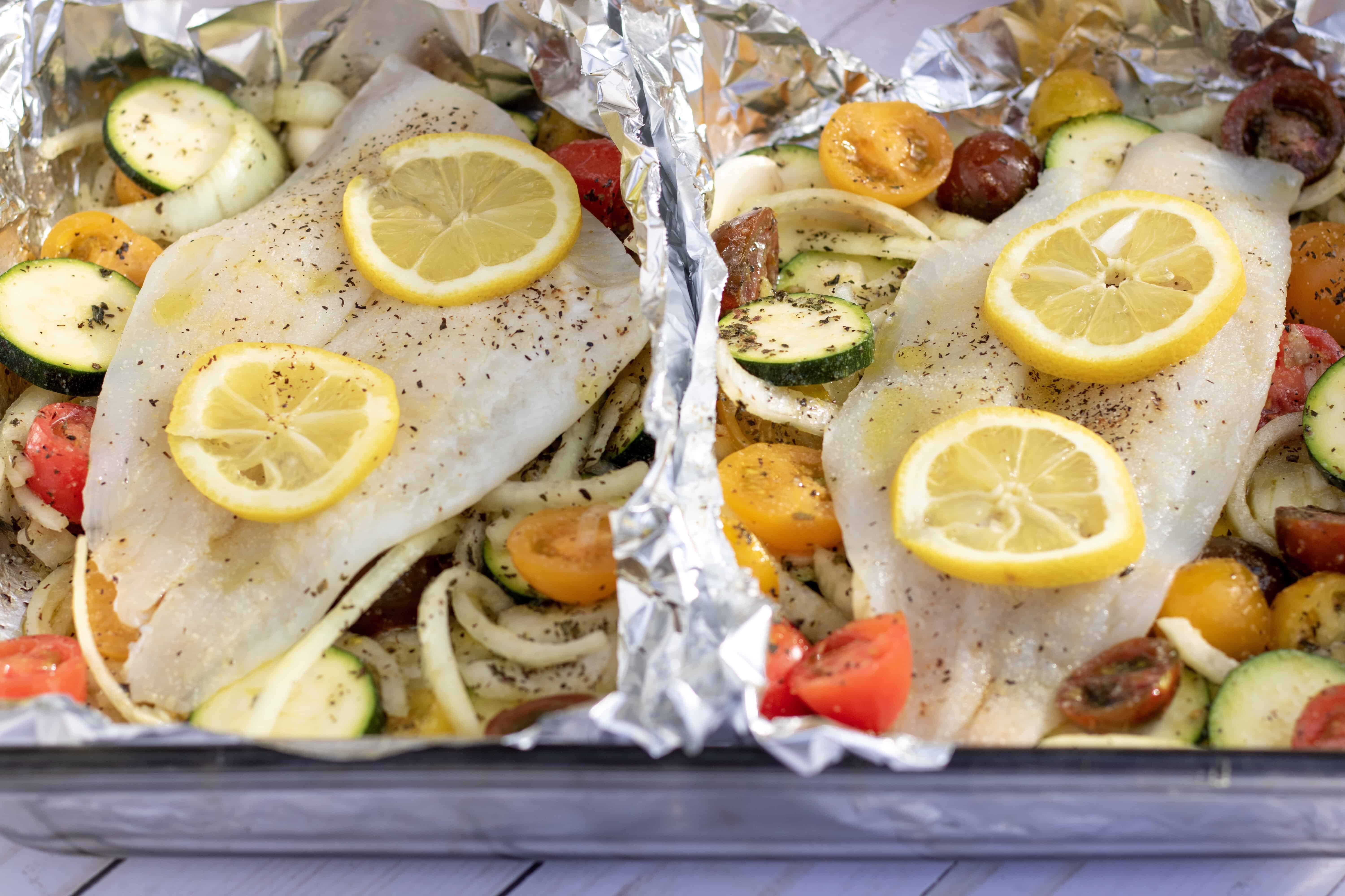 A glass 13x9” baking dish with two foil packets that are filled with vegetables and topped with white fish. It’s to be baked in the oven for an easy family dinner that’s healthy and quick to make. It’s got vibrant colorful cherry tomatoes, sliced zucchini and onions. You can see the fish is drizzled with olive oil and sprinkled with salt and pepper.