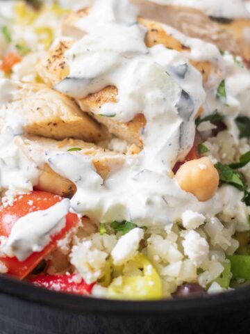 A bowl of cauliflower rice packed with Greek chicken, peppers, onions, chickpeas, tomatoes and cucumbers and topped with tzatziki sauce. A healthy meal prep recipe idea.