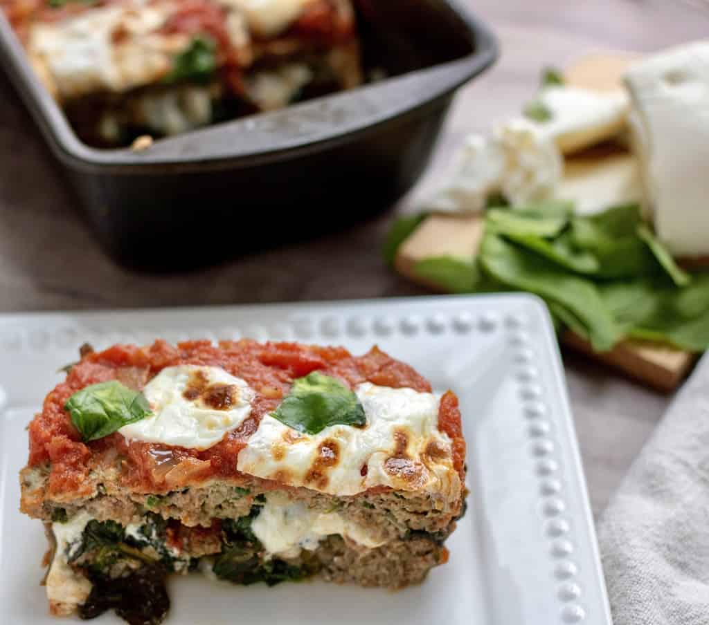 A white rectangle dish with a piece of Italian meatloaf. It’s stuffed with sautéed spinach and fresh mozzarella that’s melted. It’s topped with marinara sauce, fresh mozzarella slices and fresh basil. A loaf pan of the meatloaf is sitting in the background. It’s a quick and easy stuffed meatloaf recipe, perfect for family dinner. A small cutting board is in the background with fresh mozzarella and basil leaves