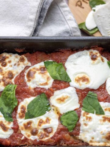 A loaf pan of Italian meatloaf that’s topped with marinara and fresh mozzarella slices that are melted and slightly golden. It’s finished with fresh basil slices.