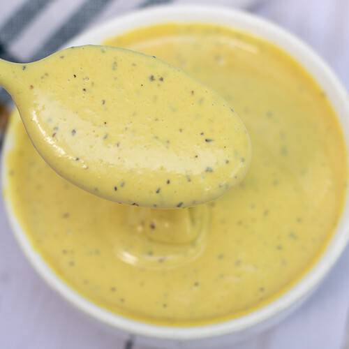 A small white bowl of maple mayo mustard sauce with a spoonful of sauce dripping into the bowl.