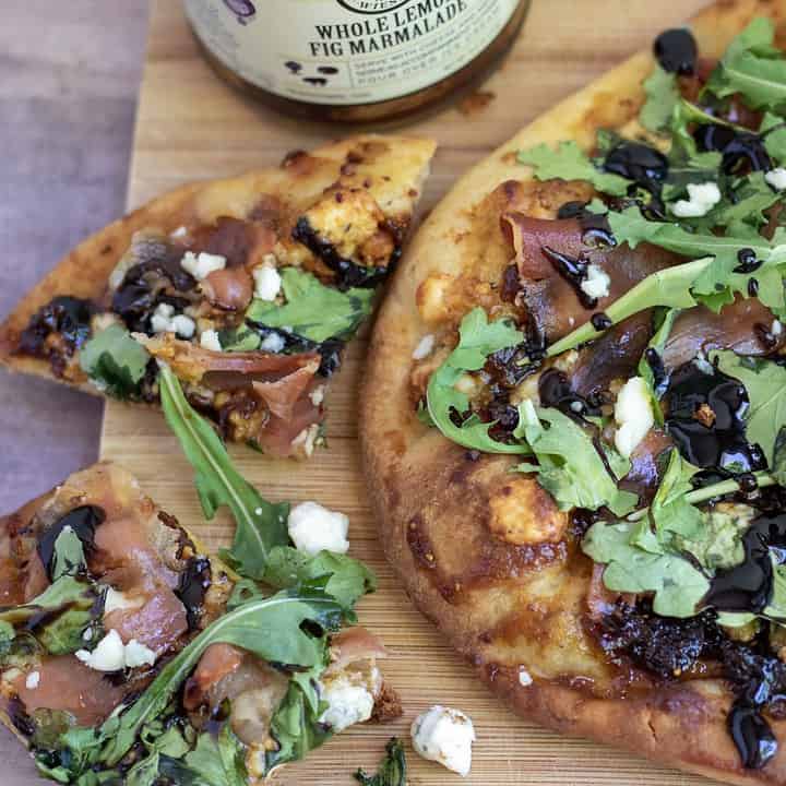 A golden crispy flatbread pizza with fig Jam, arugula, prosciutto and blue cheese crumbles. It’s finished with a balsamic drizzle. A couple pizza of cut up pizza are laying next to it with the balsamic dripping and cheese crumbles on the table.