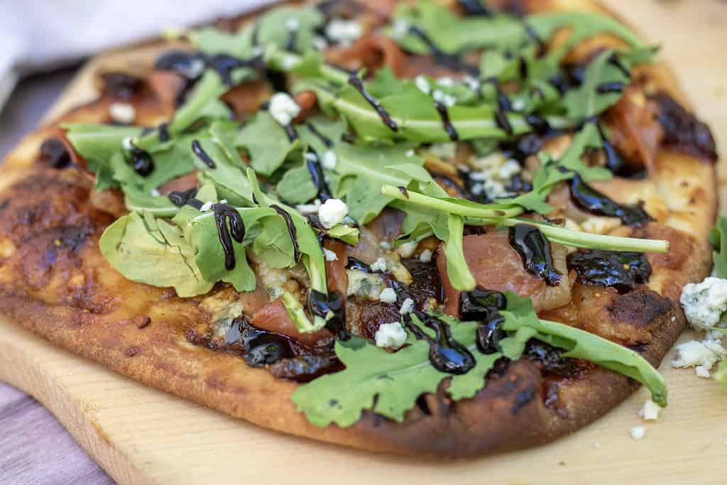 A side shot of flatbread pizza that’s sitting on a pizza paddle. You can see the fresh arugula and blue cheese crumbles. It’s got a golden crispy crust and is drizzle with balsamic