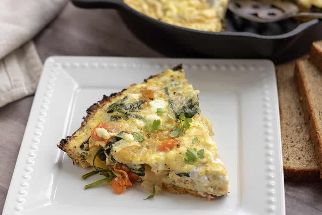 A square white dish with a slice of quiche that has a sweet potato crust, spinach, tomatoes and goat cheese. A cast iron skillet is in the background with a couple slices of whole grain bread