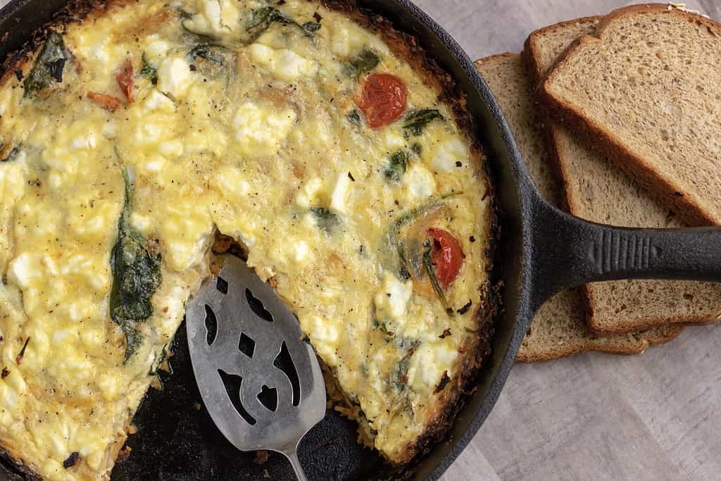 A black cast iron pan filled with quiche. It’s got a slice missing with a pie serving utensil. You can see the roasted tomatoes, goat cheese & spinach in the quiche. A couple slices of whole grain bread sit next to it.