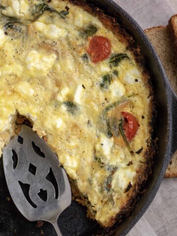 A black cast iron pan filled with quiche. It’s got a slice missing with a pie serving utensil. You can see the roasted tomatoes, goat cheese & spinach in the quiche. A couple slices of whole grain bread sit next to it.