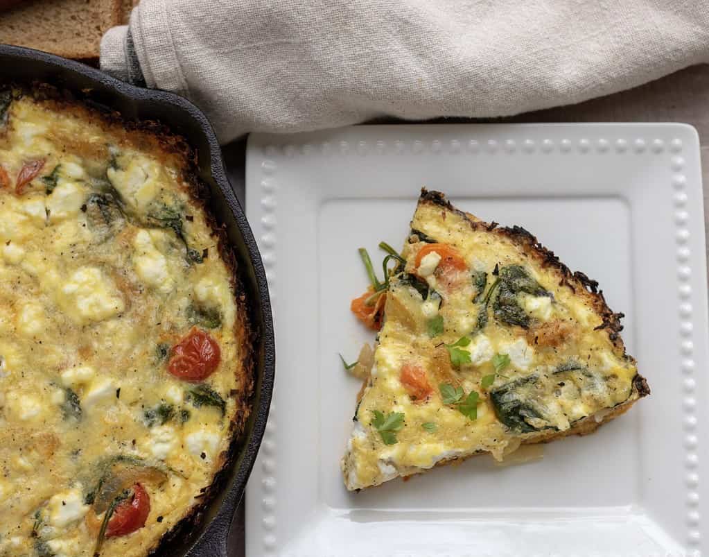 A part of a cast iron pan with quiche. There’s a white dish next to it with a slice of quiche with roasted tomatoes, caramelized onions and spinach.