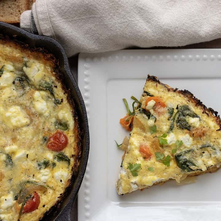 A part of a cast iron pan with quiche. There’s a white dish next to it with a slice of quiche with roasted tomatoes, caramelized onions and spinach.