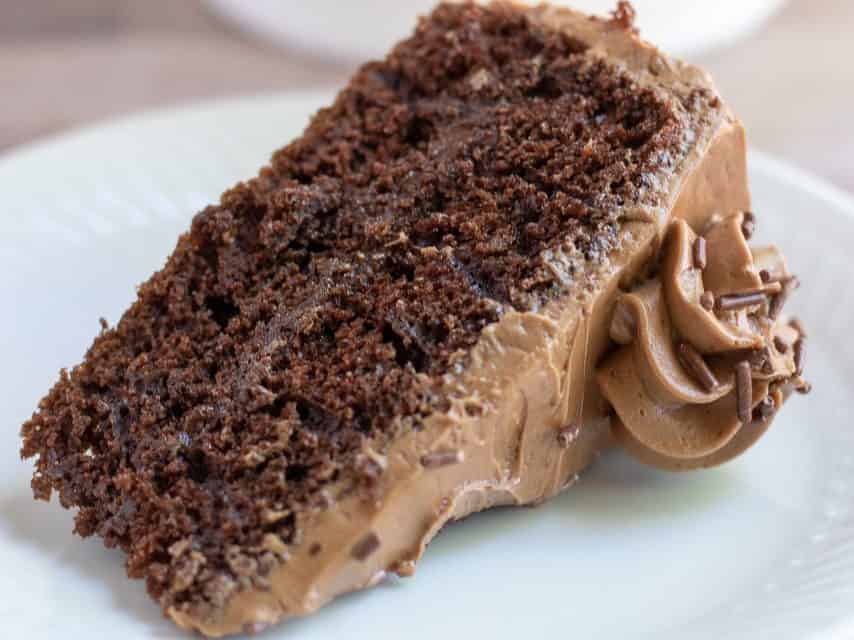 A single slice of chocolate cake on a small white dish. It's filled with a layer of creamy ganache and it's frosted with mocha swiss meringue buttercream. It's decorated with some chocolate sprinkles.
