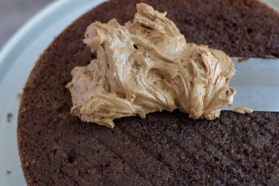 A layer of chocolate cake with a spatula smearing mocha swiss meringue buttercream on top. It's fluffy and smooth.