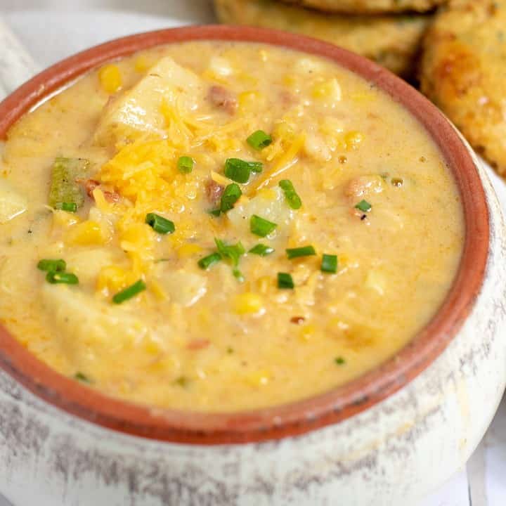 a white crock filled with creamy summer corn chowder. It’s topped with cheddar cheese and chives and loaded with zucchini and potatoes. There’s a couple of cheddar herb biscuits in the background.