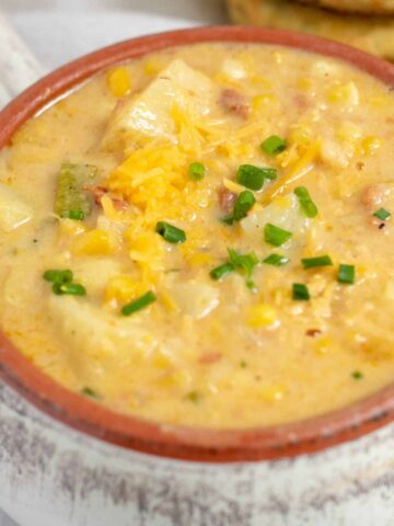 a white crock filled with creamy summer corn chowder. It’s topped with cheddar cheese and chives and loaded with zucchini and potatoes. There’s a couple of cheddar herb biscuits in the background.