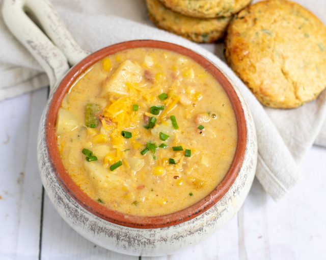 a white crock filled with creamy summer corn chowder. It’s topped with cheddar cheese and chives and loaded with zucchini and potatoes. There’s a couple of cheddar herb biscuits in the background.