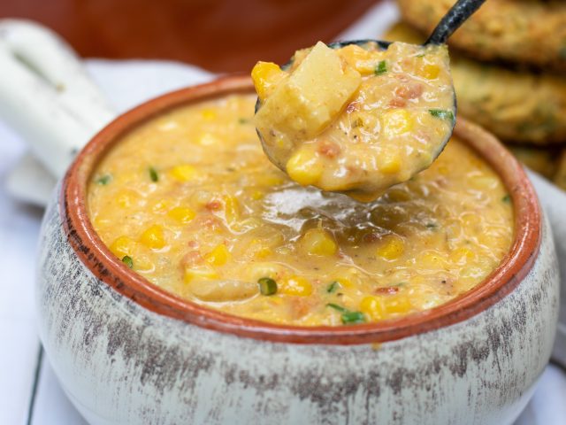 A bowl full of fresh corn chowder with a spoonful taken out of it. The spoon has chunks of yellow potatoes and zucchini and the chowder is thick and creamy.