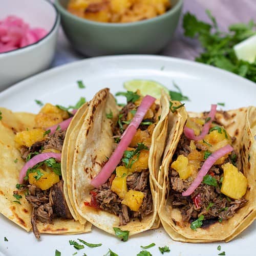 A round white plate with 3 corn tortillas that are filled with shredded beef, pineapple, pickled red onion and fresh cilantro. There's some fresh chopped cilantro on the plate and a wedge of lime. A small bowl of caramelized pineapples and another small bowl of pink pickled red onions are in the background.