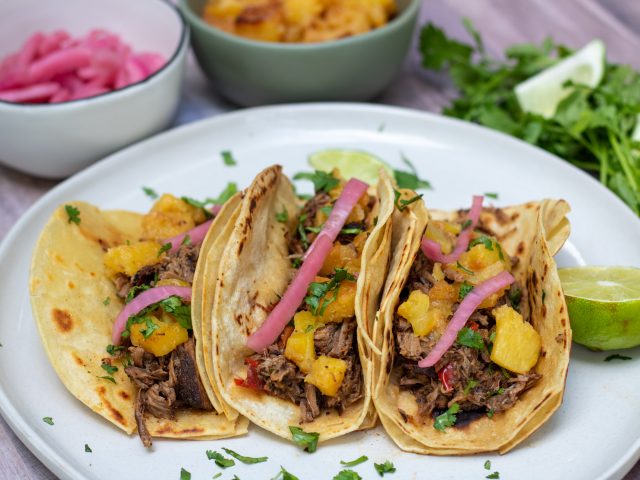 A round white plate with 3 corn tortillas that are filled with shredded beef, pineapple, pickled red onion and fresh cilantro. There's some fresh chopped cilantro on the plate and a wedge of lime. A small bowl of caramelized pineapples and another small bowl of pink pickled red onions are in the background.