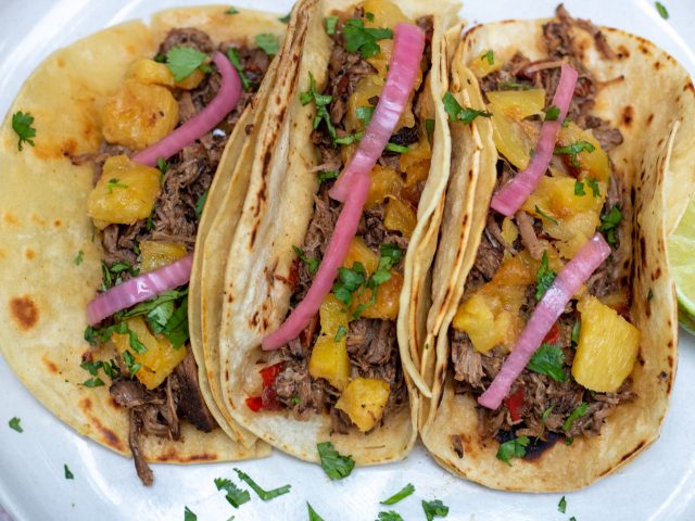 3 corn tortillas on a plate filled with crockpot chipotle barbacoa beef and topped with pink pickled red onions, fresh cilantro and caramelized pineapple.