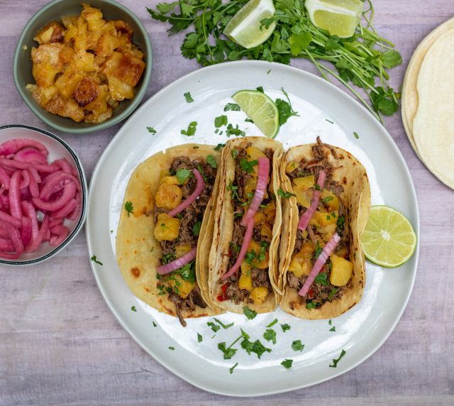 An overhead shot of a plate with 3 barbacoa tacos. They're topped with pickled red onions, pineapple and fresh cilantro. A wedge of lime is served next to the tacos and there are two small bowls, one with pickled red onions and the other with caramelized pineapple pieces. Corn tortillas are stacked in the background and a bunch of fresh cilantro.