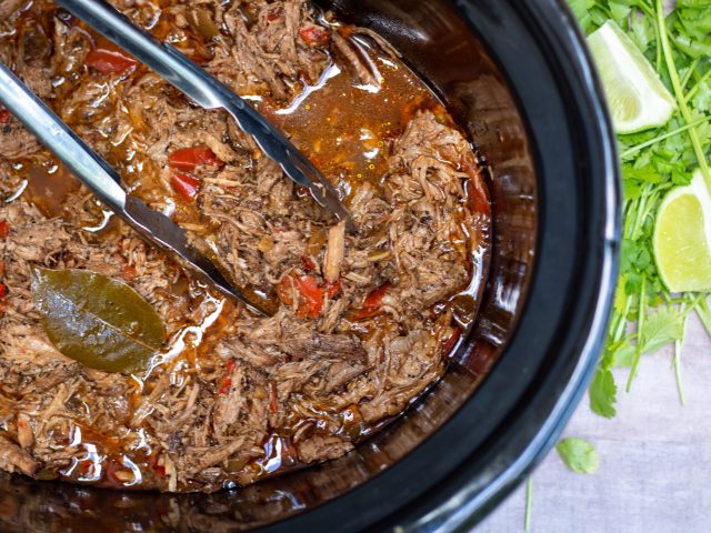 a crockpot of Mexican shredded meat that’s juicy and flavorful. There’s peppers and onions and a bay leaf. A bunch of fresh cilantro and lime is next to the crockpot.