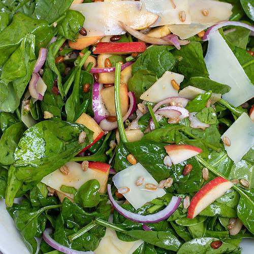 A large white bowl of spinach salad with farro, pine nuts, shaved parmesan, sliced apples, red onions and a honey dijon dressing.