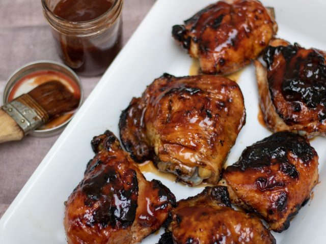 a white rectangle platter with grilled chicken that are lathered in BBQ sauce. The skin is crispy and slightly charred and a mason jar of BBQ is in the background with a pastry brush.