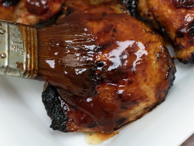 a bone-in chicken thigh with skin that’s being brushed with apricot bourbon bbq sauce. You can see the sauce dripping down the chicken. The skin is slightly blackened and crispy.