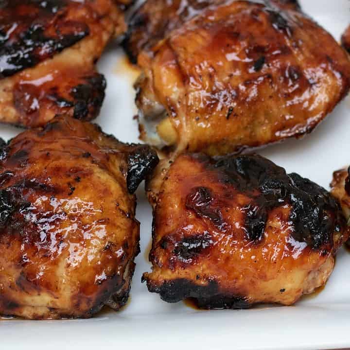 A large white rectangle platter with 6 grilled chicken thighs that have been brushed with apricot bourbon bbq sauce. The skin is crispy and slightly blackened. You can see that they're sticky and moist.