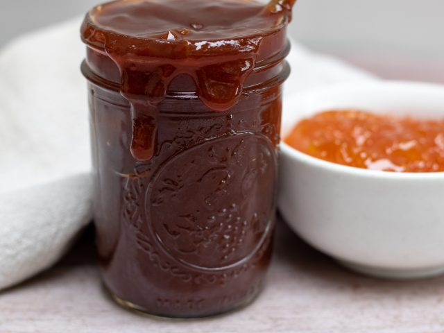 a small mason jar filled with homemade apricot bourbon bbq sauce that’s dripping down the sides. A small bowl of apricot jam is in the background.