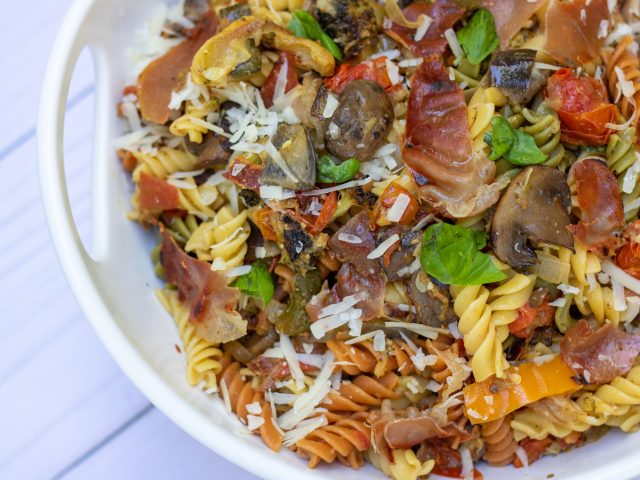 A white serving bowl filled with tri colored rotini pasta that’s tossed with grilled summer vegetables, crispy prosciutto, fresh basil and parmesan cheese. You can see the roasted tomatoes, yellow squash and broccoli in the pasta.