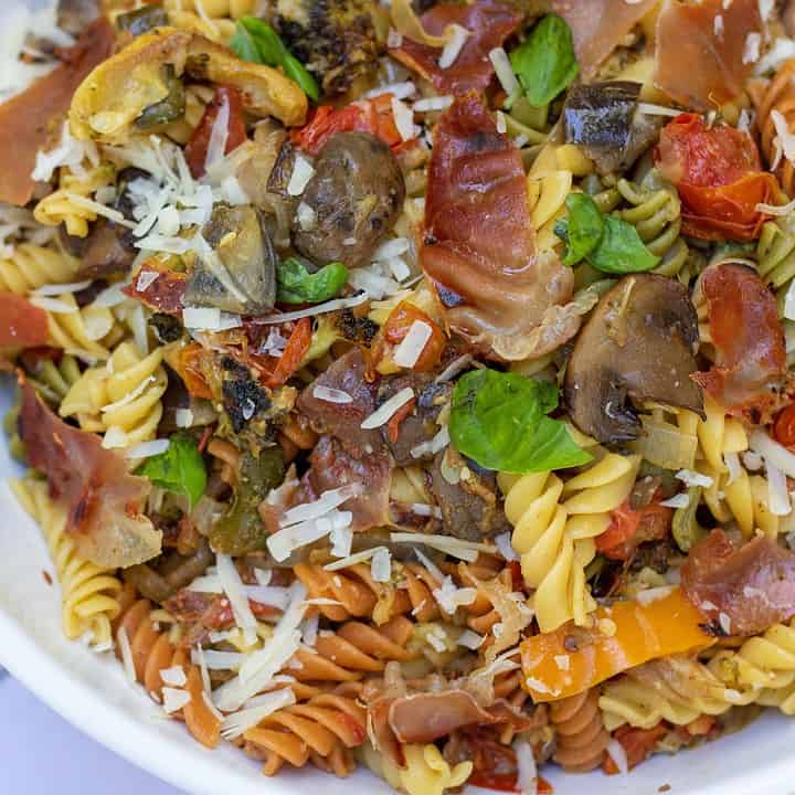 A white serving bowl filled with tri colored rotini pasta that’s tossed with grilled summer vegetables, crispy prosciutto, fresh basil and parmesan cheese. You can see the roasted tomatoes, yellow squash and broccoli in the pasta.