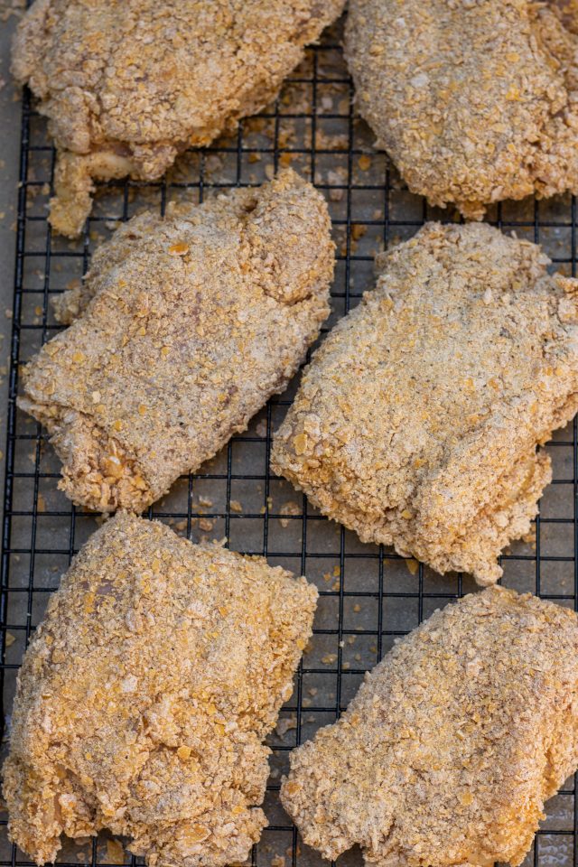 6 pieces of chicken thighs that are laid on a cooling rack lined baking sheet. They're coated in crushed corn flakes and ready to be baked in the oven. You can see the crumbs from the coating on the baking sheet.
