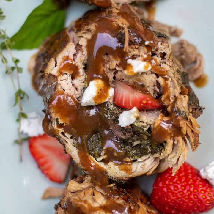 Pork tenderloin that’s stuffed with sautéed spinach, sliced strawberries, and goat cheese has been sliced and drizzled with balsamic gravy. It’s served on a light green serving platter and there’s fresh strawberry slices and goat cheese on the platter