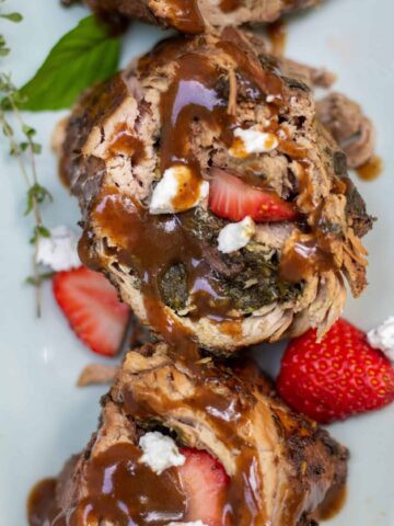 Pork tenderloin that’s stuffed with sautéed spinach, sliced strawberries, and goat cheese has been sliced and drizzled with balsamic gravy. It’s served on a light green serving platter and there’s fresh strawberry slices and goat cheese on the platter