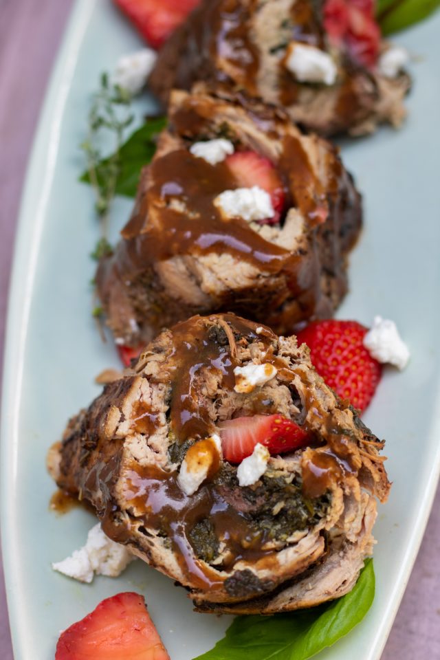 a serving plate with slices of pork tenderloin that have been stuffed with spinach, strawberries and goat cheese. You can see the fresh strawberry slices on the plate along with fresh basil, and crumbles of goat cheese. The pork is drizzled with a golden brown balsamic honey gravy.
