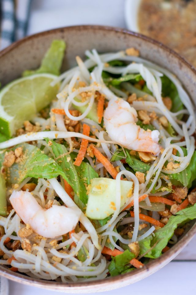 A brown speckles bowl with rice noodles, chopped lettuce, shredded carrots, cucumber, bean sprouts, basil and mint. It's topped with shrimp and a wedge of lime.