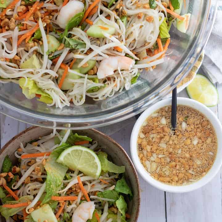 A large glass bowl of spring roll salad. There's a smaller bowl in the front with rice noodles, carrots, bean sprouts, cucumber, fresh mint and basil. It's topped with shrimp and crushed peanuts. Next to the bowls is a smaller bowl with peanut dressing with a small spoon in it.