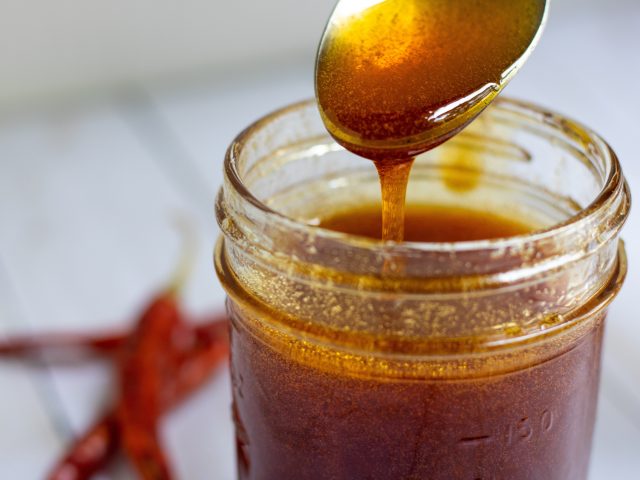 A mason jar of hot honey with a spoonful being taken out of it that's dripping into the jar. You can see some dried chili peppers in the background.