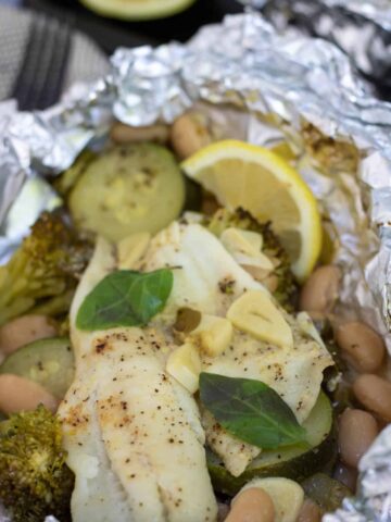 A foil packet filled with zucchini, broccoli, white beans and white fish. There's slices of garlic, fresh basil and lemon on top.