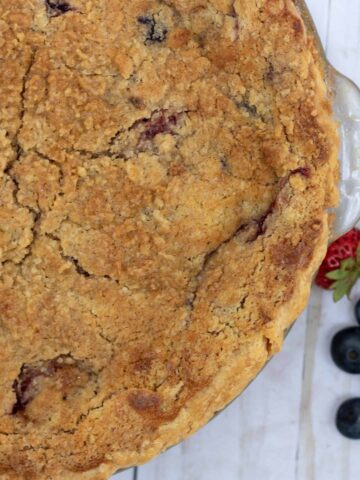 A berry pie with crumb topping. It's made with fresh summer berries like blackberries, blueberries and strawberries. The crumb topping is golden brown. There are a couple of fresh blueberries and strawberries next to the glass pie plate.