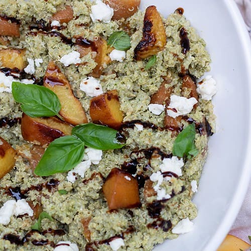 A white serving bowl with pesto quinoa, grilled peaches, goat cheese crumbles and balsamic drizzle. It's garnished with fresh basil leaves. There's a couple of fresh peach slices and fresh thyme sprigs next to the bowl in the background.