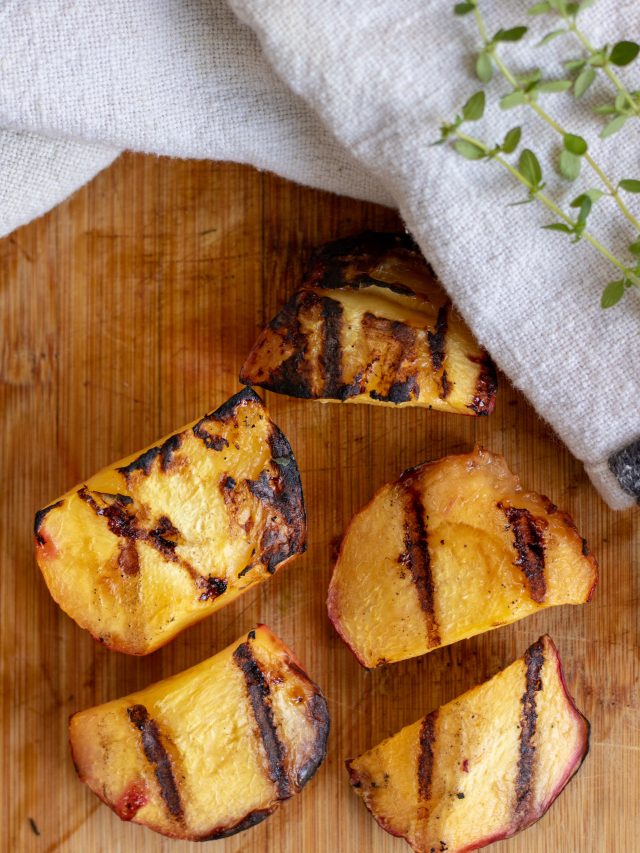 A wooden cutting board with pieces of grilled peaches. They have perfect grill marks and there's a sprig of fresh thyme next to them.